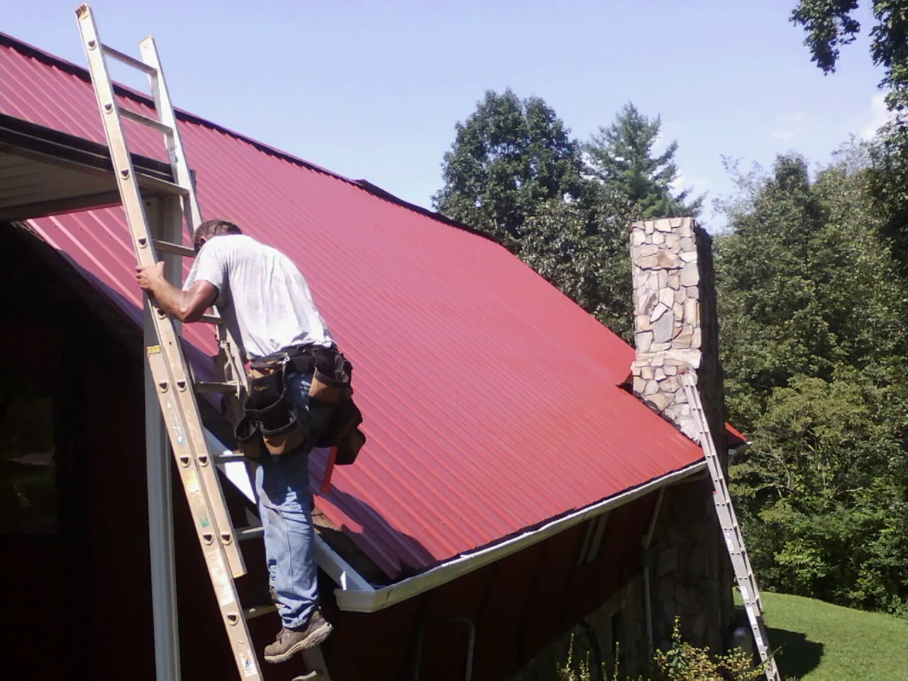 Roofer on a ladder beside a red metal roof and stone chimney — Soffit & Fascia Repair by Roofing Brothers in Toccoa